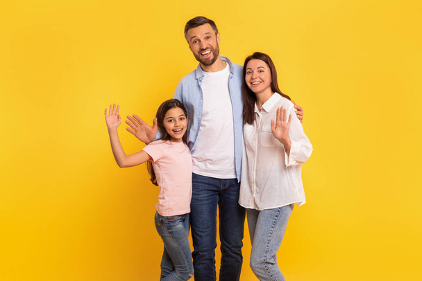Hello. Cheerful family of parents and daughter kid waving hands smiling at camera, hugging over yellow studio background. Man embraces his wife and child, gesturing hi greeting together - Photo, Image