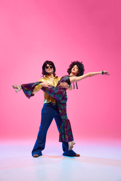 Динамічний портрет танцюристів дуету, чоловіка і жінки, що позують у ретро модному одязі на фоні градієнтної рожевої студії. Концепція американської культури, 1970-х, 1980-х років мода, музика, порівняння епох. - Фото, зображення
