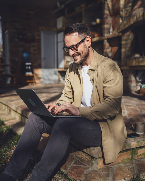 Egy férfi kaukázusi modern férfi ül az erkélyen terasz egyedül otthon viselet ing munka laptop számítógép nappal távoli boldog mosoly magabiztos siker koncepció másolás tér - Fotó, kép