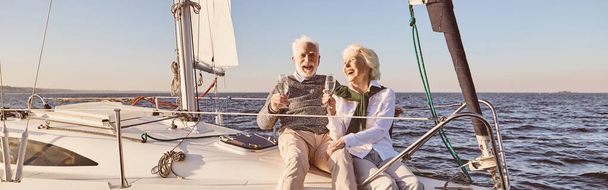 Ευτυχισμένο ζευγάρι ηλικιωμένων που κάθονται στην πλευρά του ιστιοφόρου ή κατάστρωμα γιοτ επιπλέουν στη θάλασσα. Άντρας και γυναίκα πίνουν κρασί ή σαμπάνια και γελούν. Ιστιοπλοΐα, ταξίδια και οι άνθρωποι έννοια. Οριζόντια βολή - Φωτογραφία, εικόνα