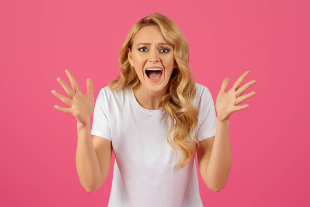 Boos trieste jonge blonde vrouw schreeuwend hand in hand, flipt over roze studio achtergrond. Mensen emoties, reactie op slecht nieuws op het werk, ruzie en stress concept - Foto, afbeelding