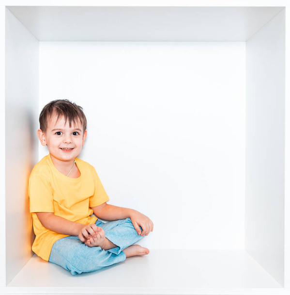 Um menino bonito em uma camiseta amarela e calças azuis senta-se em um nicho branco na mobília em seu quarto. Bebê em uma caixa branca quadrada. - Foto, Imagem