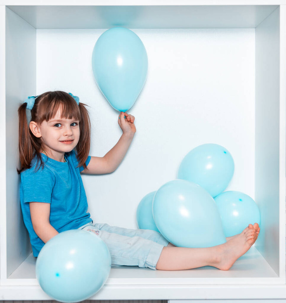 Egy csinos kislány kék pólóban és kék nadrágban ül egy fehér bútorrésben a szobájában. Baba egy négyzet alakú fehér dobozban. Lány kék lufikkal. - Fotó, kép