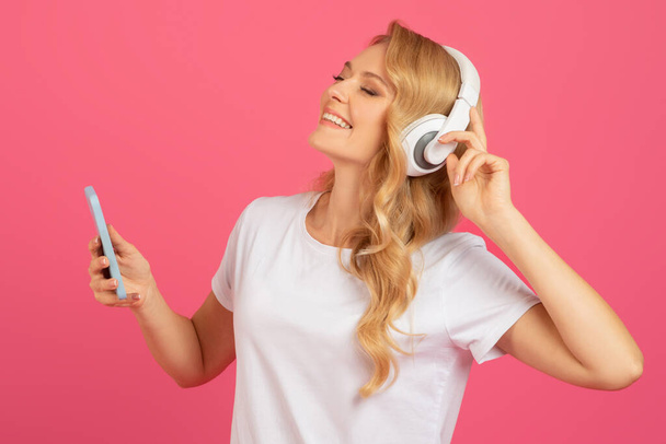 Портрет улыбающейся блондинки в белых футболках и с натянутыми наушниками, наслаждающейся музыкой в Интернете на розовом студийном фоне. Леди с удовольствием слушает любимые песни - Фото, изображение