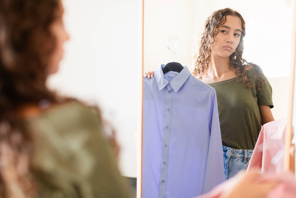 Problem of fashion choice. Unhappy European teenager girl facing shopping issue, displaying discontent while choosing clothes, standing with two shirts near mirror indoors. Selective focus - Photo, Image