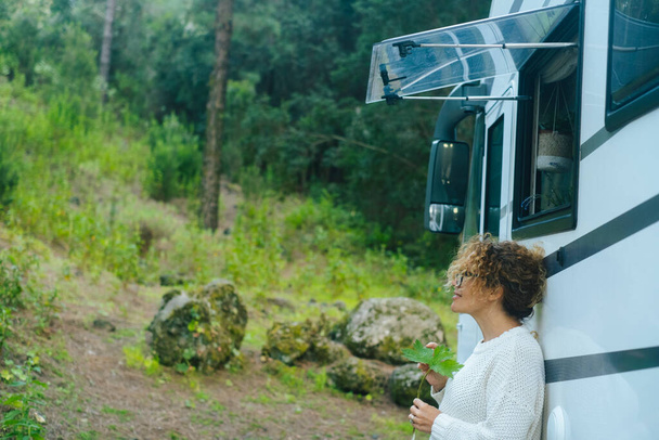 Büyük bir karavan karavanı karavanına karşı duran bir kadın yeşil ormandaki gezici vanLife durağında doğa ve huzur dolu bir günün tadını çıkarıyor. İnsanlar ve seyahat yaşam tarzı ya da tatil minibüsü kiralama - Fotoğraf, Görsel