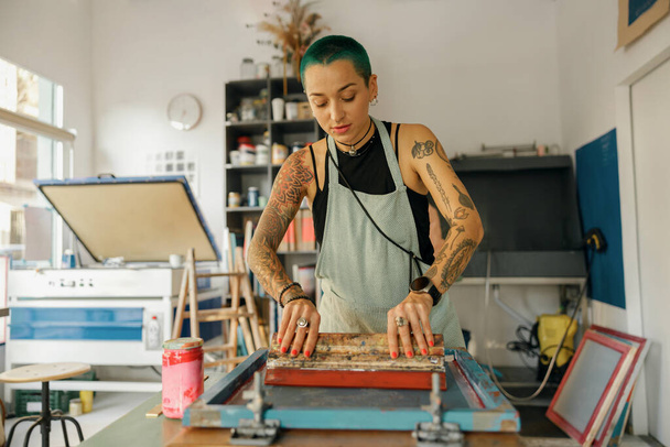 Vrouwelijke werknemer met squeegee drukt inkt op frame werken in design studio. Zeefdruk - Foto, afbeelding