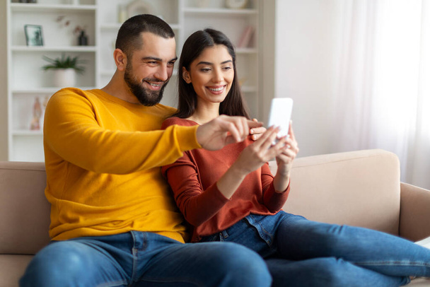 Onnellinen nuori mies ja nainen käyttävät älypuhelinta rentoutuessaan sohvalla yhdessä, naimisissa oleva valkoihoinen pari halailee ja katselee matkapuhelinnäyttöä, hymyilevät puolisot käyttävät uusinta mobiilisovellusta - Valokuva, kuva