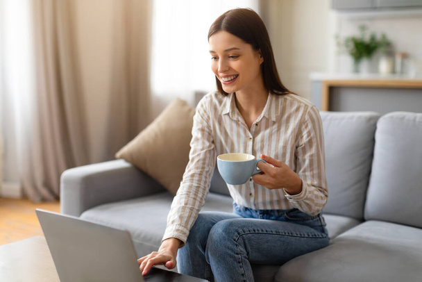 Iloinen nuori nainen nauttii kuppi kahvia, kun käytät hänen kannettava tietokone, mukavasti istuu sohvalla viihtyisä ja hyvin valaistu olohuone sisustus, nainen selailee internetiä tai työskentelee etänä - Valokuva, kuva