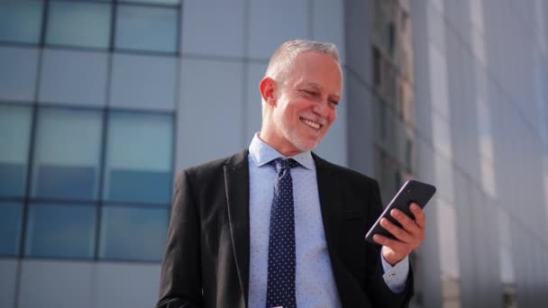 Retrato de um homem de negócios maduro em um blazer, gravata e camisa de vestido, de pé na frente de um edifício, está sorrindo enquanto segura um celular. Empresa ceo sênior usando um telefone inteligente no local de trabalho. Alto - Filmagem, Vídeo