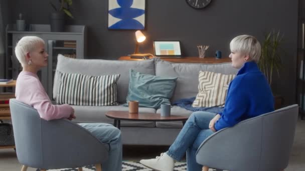 Volledig shot van twee jonge blanke vrouw communiceren met elkaar met behulp van de hand vinger gebarentaal, zitten in zachte grijze fauteuils in modern appartement - Video