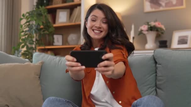 Μια ενήλικη καυκάσια γυναίκα κάθεται στο σπίτι χαρούμενο χαμόγελο παίζουν παιχνίδια βίντεο ψυχαγωγική δραστηριότητα έχοντας διασκέδαση κρατήστε το κινητό τηλέφωνο χρήση smartphone διασκεδάστε online - Πλάνα, βίντεο