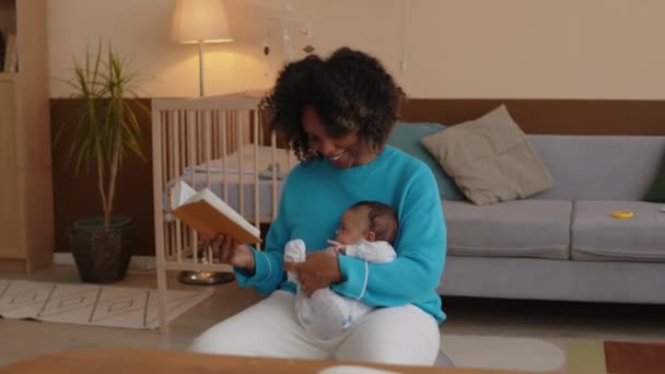 Media toma de la joven mujer negra sosteniendo y balanceando al bebé mientras se sienta en la bola en forma en la sala de estar y libro de lectura - Metraje, vídeo