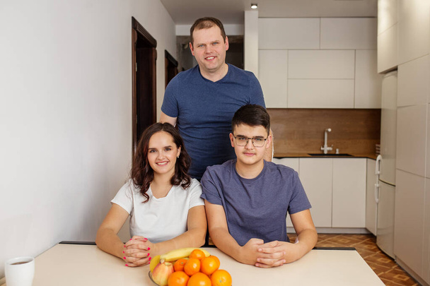 Szczęśliwa rodzina trojga siedzi w dobrze oświetlonej nowoczesnej kuchni ze świeżymi owocami na stole - Zdjęcie, obraz