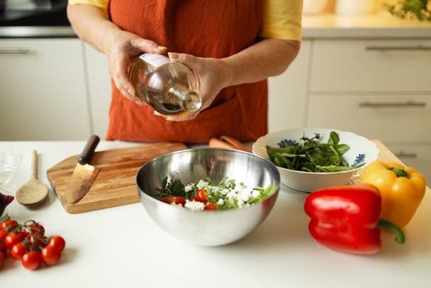 Женщина приправляет овощной салат во время приготовления пищи на кухне. Неузнаваемый шеф-повар добавляет оливковое масло во время приготовления вегетарианской еды дома. Концепция здорового питания.  - Фото, изображение
