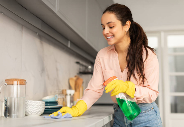 Lächelnde Frau in Bluse und gelben Handschuhen, fröhlich die Küchenarbeitsplatte mit blauem Tuch und grüner Sprühflasche in der Hand wischend, freier Raum - Foto, Bild