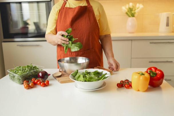 Senior 60s rubia caucásica sonriente mujer sosteniendo un cuchillo y espinacas frescas enteras preparando un tazón de ensalada de dieta saludable en su cocina ecológica rústica. Foto de alta calidad - Foto, imagen