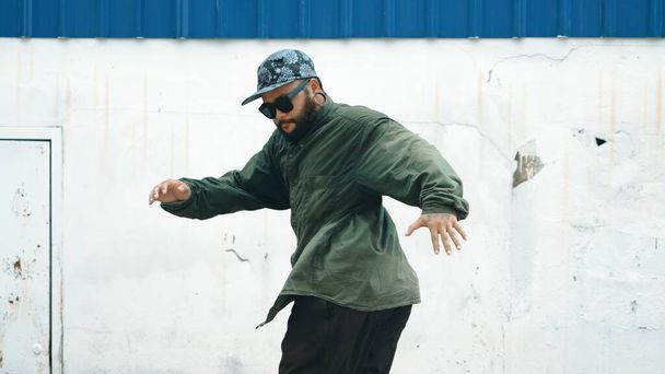 Hipster tančící b-boy krok na ulici s modrou zdí. Asijské přestávka tanečník praxe street dance zatímco strečové paže. Hip hop choreograf předvádí energický tanec. Venkovní sport2024. Endeavor. - Fotografie, Obrázek