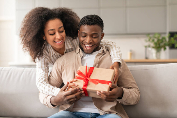 Genç, neşeli siyah çift bayramları ve özel günleri kutluyor. Kocasına hediye paketini sunan kadın evdeki koltukta oturup keyif paylaşıyor. Sevgililer Günü hediyesi - Fotoğraf, Görsel