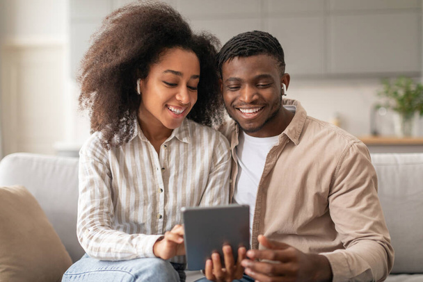 Uśmiechnięta afro-amerykańska para z bezprzewodowymi słuchawkami wykorzystująca cyfrowy tablet do aktywności online i zabawy, zanurzona w technologii gadżetów, ciesząca się przytulnym weekendem na kanapie w salonie - Zdjęcie, obraz