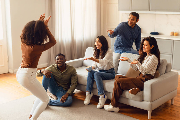 Diverse group of friends engages in game of charades, enjoy guessing and acting in cozy living room at home, creating carefree atmosphere of joy and camaraderie during their weekend celebration - Photo, Image