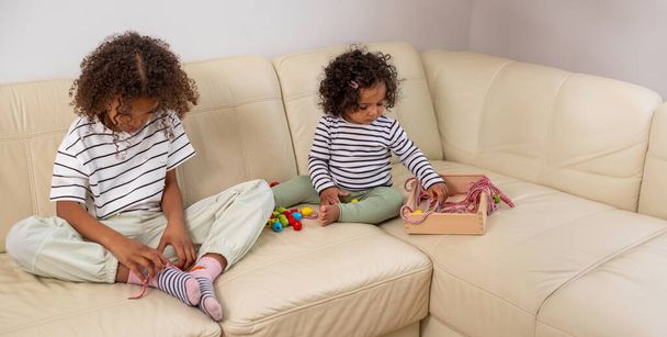 Baner dwóch czarnych dziewczyn grających na kanapie z zabawkami edukacyjnymi. Zarówno skoncentrowane, oparte na rozwoju zabawki, symbolizujące zajęcie i zainteresowanie nauką. Wysokiej jakości zdjęcie - Zdjęcie, obraz
