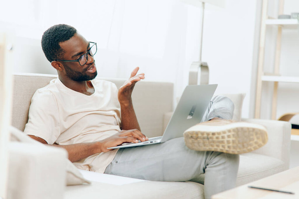 Freelancer Afro-Americano Trabalhando em Laptop em um Home Office Moderno, Sorrindo e Envolvido no Ciberespaço Educação Um jovem negro é visto sentado em um sofá confortável, digitando em seu computador - Foto, Imagem