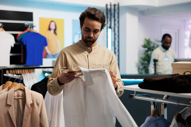 Man checking stylish casual shirt in shopping mall department outlet. Young customer examining trendy garment on hanger while choosing apparel in retail center fashion boutique - Photo, Image