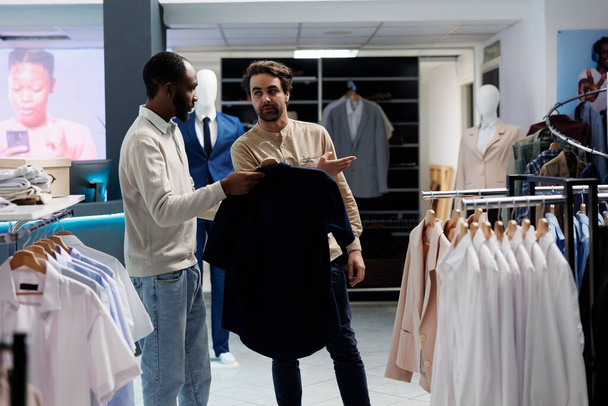Afrikanisch-amerikanischer Mann mit Hemd am Kleiderbügel und fragt Mitarbeiter eines Einkaufszentrums nach Größenoptionen. Boutique-Mitarbeiterin zeigt auf neue Kleiderständer, die Kunden bei der Auswahl ihres Outfits helfen - Foto, Bild