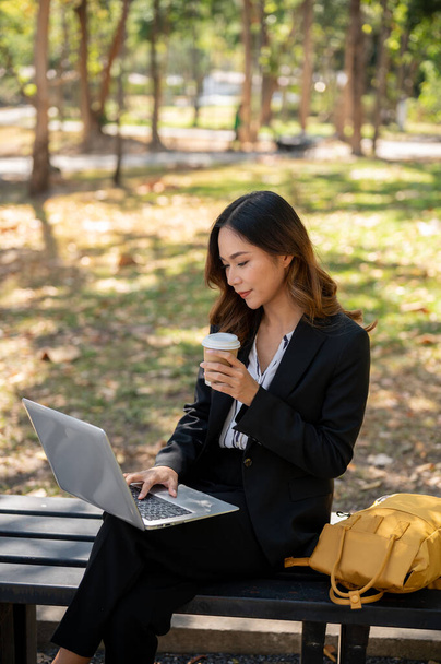 美しいアジアの実業家がコーヒーを飲んで,街の公園のベンチに座り,ラップトップコンピュータに取り組んでいる.. - 写真・画像