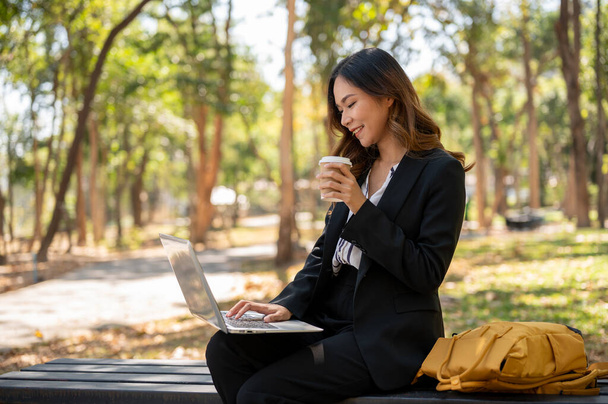 Egy elegáns ázsiai üzletasszony távirányítással dolgozik egy parkban, kávét kortyolgat és a laptopján dolgozik. modern városi élet, vállalati élet - Fotó, kép