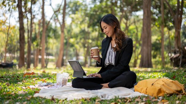 Güzel bir Asyalı iş kadını kahvesini yudumluyor ve dijital tabletini parkta bir piknik minderinde hazırlıyor.. - Fotoğraf, Görsel