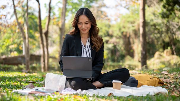 Olumlu ve çekici Asyalı bir iş kadını parkta çalışıyor, dijital tabletini piknik paspası üzerinde çalışıyor.. - Fotoğraf, Görsel