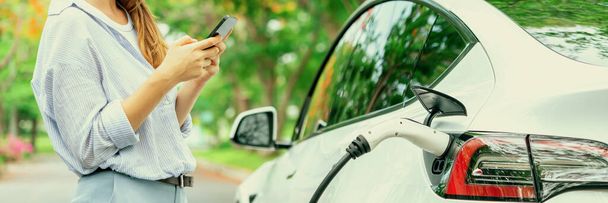 Panorama banner woman use smartphone online banking application to pay for EV car battery charging from EV charging station during autumn vacation holiday trip at national park or forest. Exalt - Photo, Image