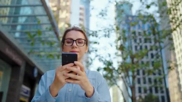 Λευκή γυναίκα με γυαλιά περπατά στην πόλη και χρησιμοποιεί smartphone. Οι ουρανοξύστες είναι στο βάθος. Επικοινωνία, εργάσιμη μέρα, πολυάσχολη έννοια της ζωής. Αργή κίνηση - Πλάνα, βίντεο
