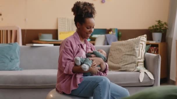 Media toma de la joven madre afroamericana saltando sobre la pelota en forma en la sala de estar mientras balancea al bebé en sus brazos - Imágenes, Vídeo