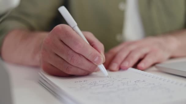 Close up of male hands taking notes on notepad using white pen at home interior. Caucasian male freelancer in casual wear handwriting document during remote work at cozy atmosphere. Report concept. - Footage, Video