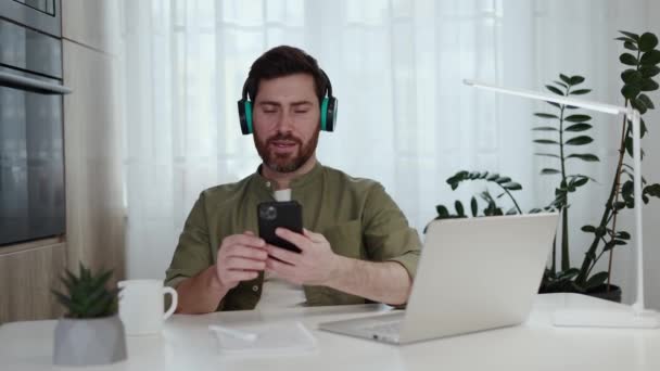 Joyful caucasian man using wireless headphones and smartphone for listening music while resting from remote work. Bearded freelancer in casual wear sitting at home office with laptop and having fun. - Footage, Video