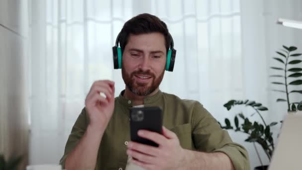 Actieve man browsen playlist op persoonlijke smartphone en het kiezen van favoriete nummer tijdens het rusten op de werkplek met draagbare laptop. Glimlachende man met draadloze hoofdtelefoon in ritme in gezellig huis. - Video