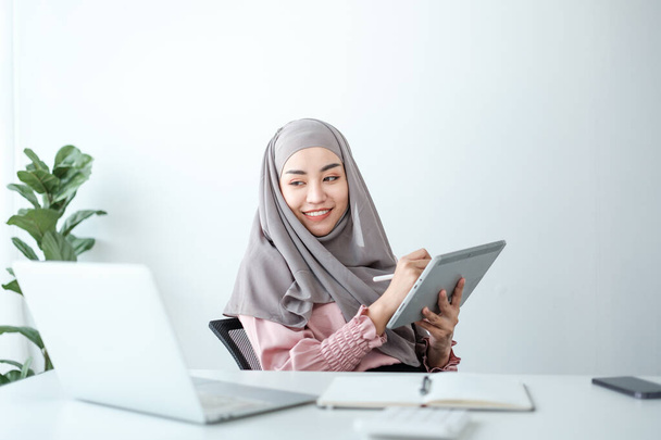 joven mujer musulmana bastante moderna en hijab trabajando en el ordenador portátil en la sala de oficina, la educación en línea, el trabajo remoto freelancer, utilizando el teléfono inteligente. Foto de alta calidad - Foto, imagen