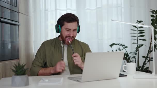 Cheerful man enjoying music through wireless headphones while sitting at home desk with modern laptop. Joyful male enthusiast in casual attire using pencils as drumsticks to play virtual drums. - Footage, Video