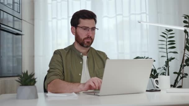 Pensive valkoihoinen mies engrossed työprosessissa ja kirjoittamalla kannettava kannettava tietokone. Huomaavainen mies, jolla on tummat hiukset, istuu pöydässä kotitoimistossa säilyttäen samalla keskittymisen ja joskus katsomalla sivuun. - Materiaali, video