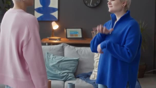 Наклоните снимок двух кавказских девушек, разговаривающих на языке жестов, стоя в современной квартире с синей абстрактной картиной на темно-серой стене на заднем плане - Кадры, видео