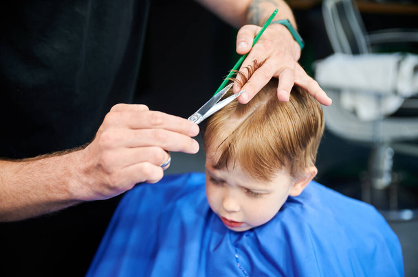 Netter Junge besorgt über neuen Haarschnitt. Liebenswerter Client mit wechselndem Stil. Zugeschnittenes Foto vom Friseur, der Jungen die Haare schneidet. - Foto, Bild