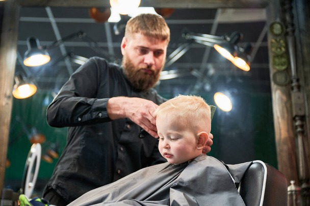 Um rapazinho giro a cortar o cabelo. Um rapaz loiro e curioso, pela primeira vez na barbearia. Cabeleireiro profissional fazendo corte de cabelo elegante por barbeador. - Foto, Imagem