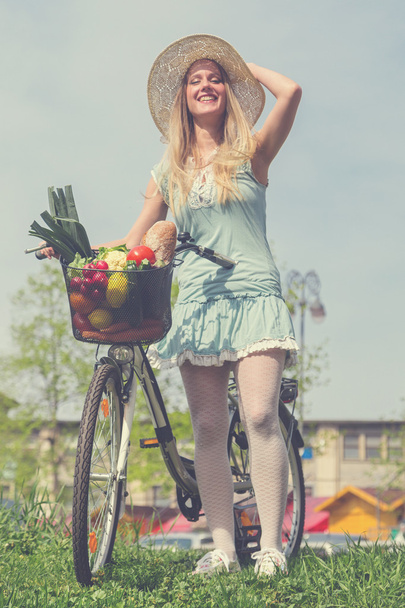 Attractive blonde woman with straw hat posing next to bike with basket  full of groceries. - Фото, зображення