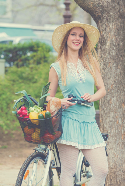 Attractive blonde woman with straw hat standing in the park and posing next to bike with basket full of groceries. - Photo, Image