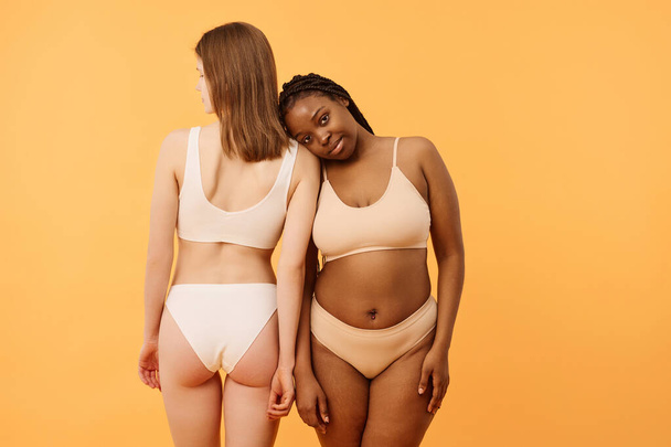 Konceptualne zdjęcie dwóch młodych, zróżnicowanych etnicznie kobiet w minimalistycznej bieliźnie, pozujących do kamery, ciepłe morelowe tło - Zdjęcie, obraz