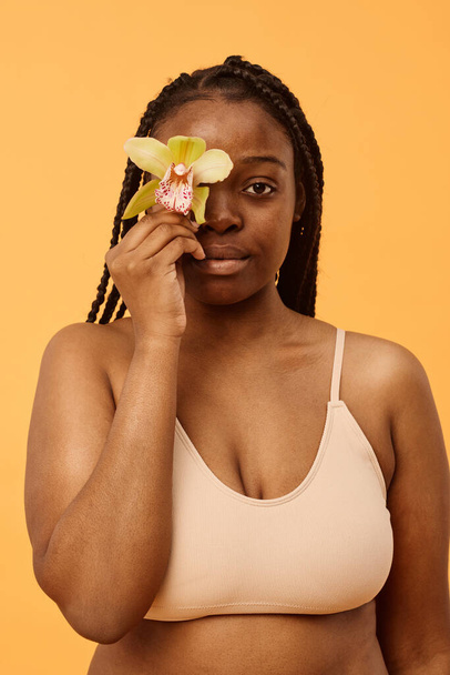 Pionowa talia w górę studio strzał młodej czarnej kobiety noszącej bralette pokrycie oka ze świeżym kwiatem storczyków patrząc na aparat - Zdjęcie, obraz