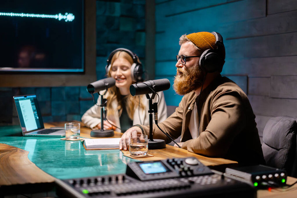 Two young stylish radio show hosts record fresh podcast episode in home loft studio apartment. Attractive energetic co-hosts discuss important topics live on air in an evening show - Photo, Image